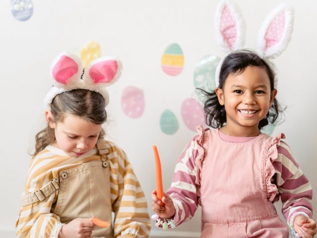Two little girls dressed in pinks and pastels wearing fluffy bunny ears. One little girl is holding a carrot and smiling