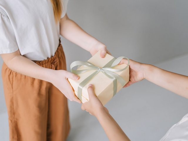A close-up of the chest down of someone handing another person a box-shaped gift. It’s wrapped in white paper with a pale green ribbon around it.