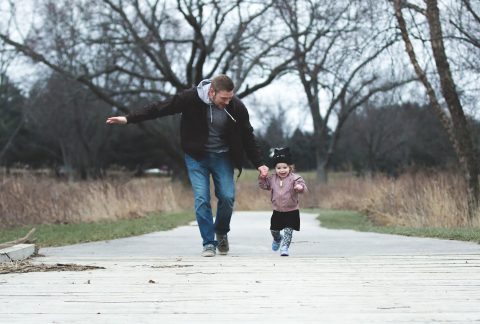 A man in a brown jacket and jeans holding hands and running in a park with a little girl in a pink coat