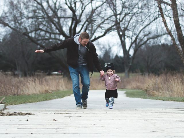 A man in a brown jacket and jeans holding hands and running in a park with a little girl in a pink coat